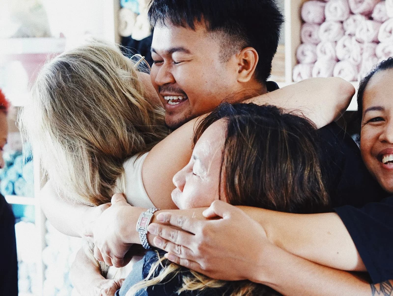 Five people crushed into one laughing group hug in a salon, a man in the middle with his eyes shut, shelves of rolled towels behind them.
